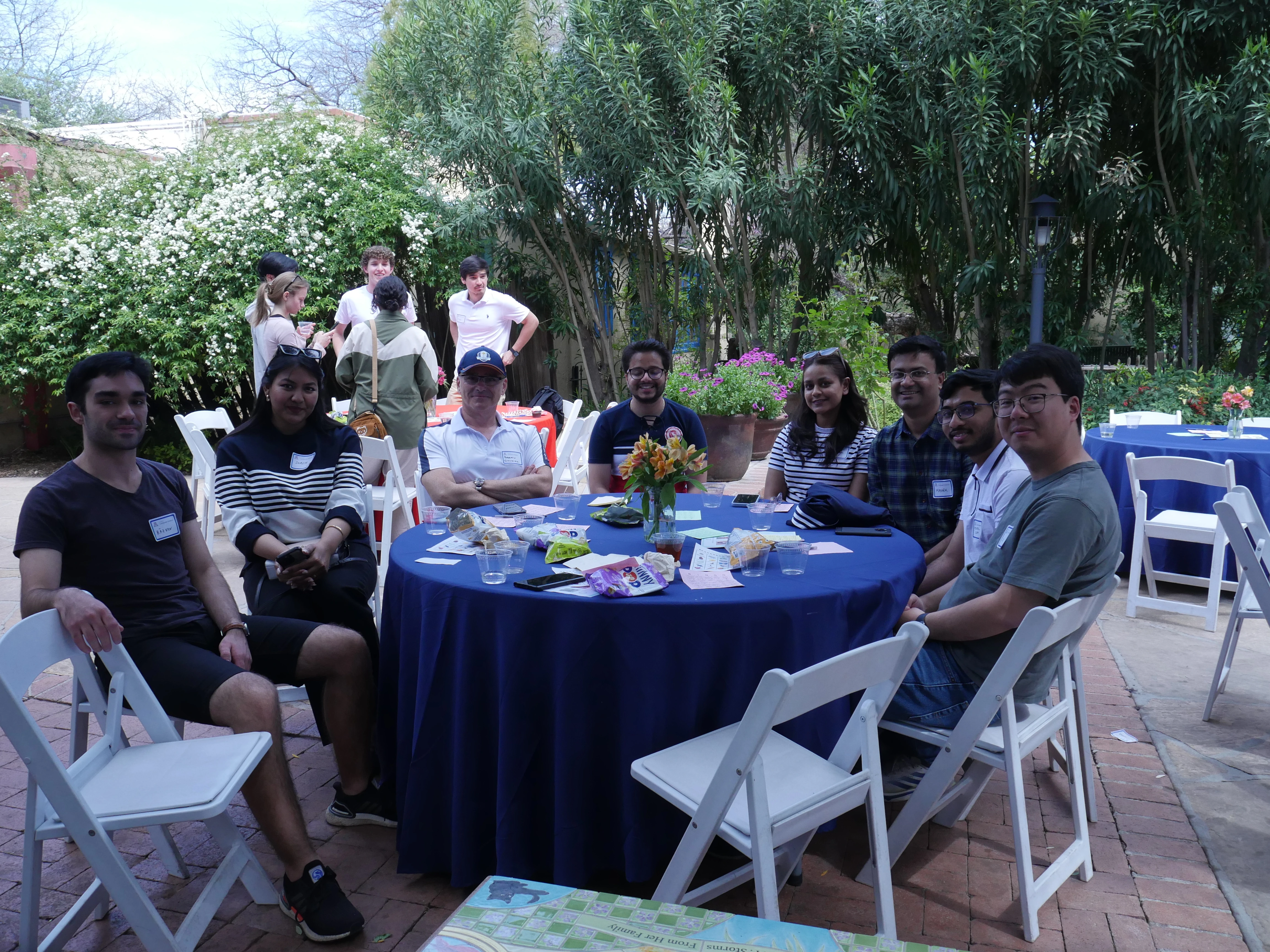 8 event attendees at a table with a blue cloth. CS ambassadors stand at a table in the background